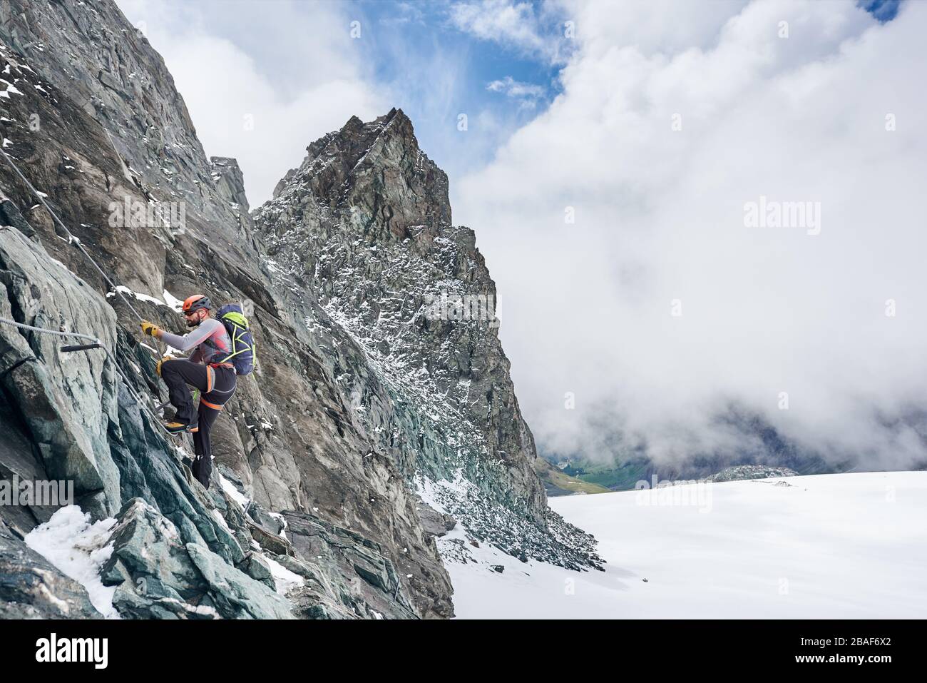 Seitenansicht des Felskletterers in Schutzhelm mit festem Seil, während er felsigen Berg mit schönem Himmel auf Hintergrund aufsteigt. Alpinist mit Rucksack Klettern natürliche Felsformation. Konzept des Alpinismus Stockfoto