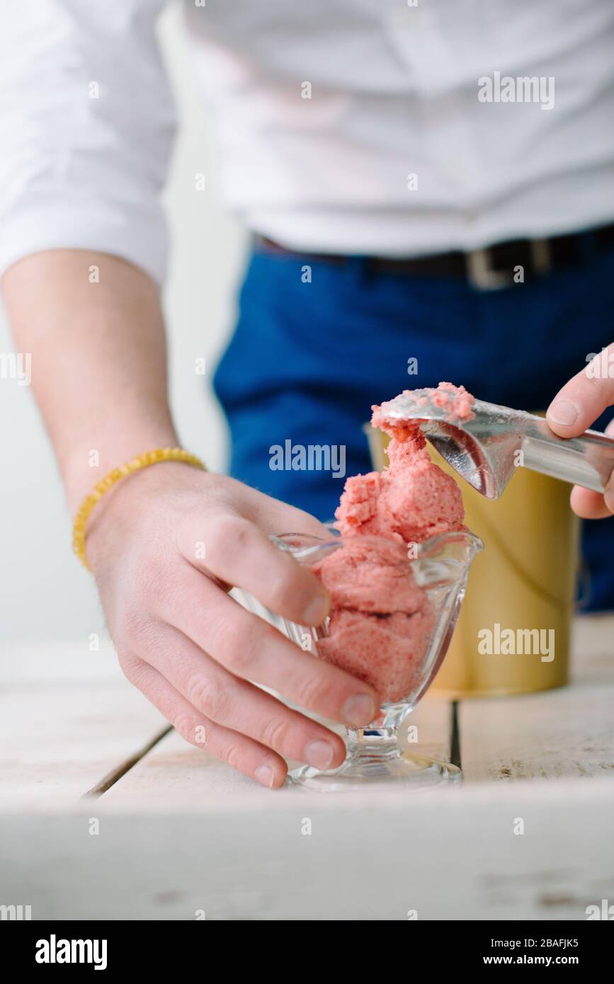 Die Hände des Mannes schaufeln rosafarbenes Eis in Tassen mit alter Schaufel auf weißem Holztisch Stockfoto