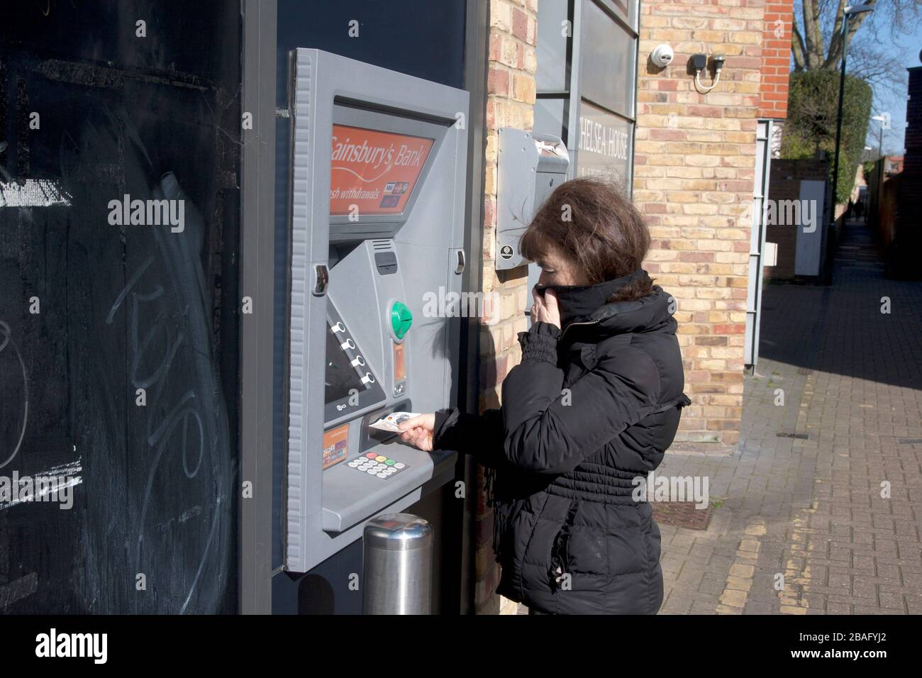 Frau mittleren Alters während Coronavirus 2020 am Geldautomaten, der ihr Gesicht bedeckt und Bargeld einsammelt Stockfoto