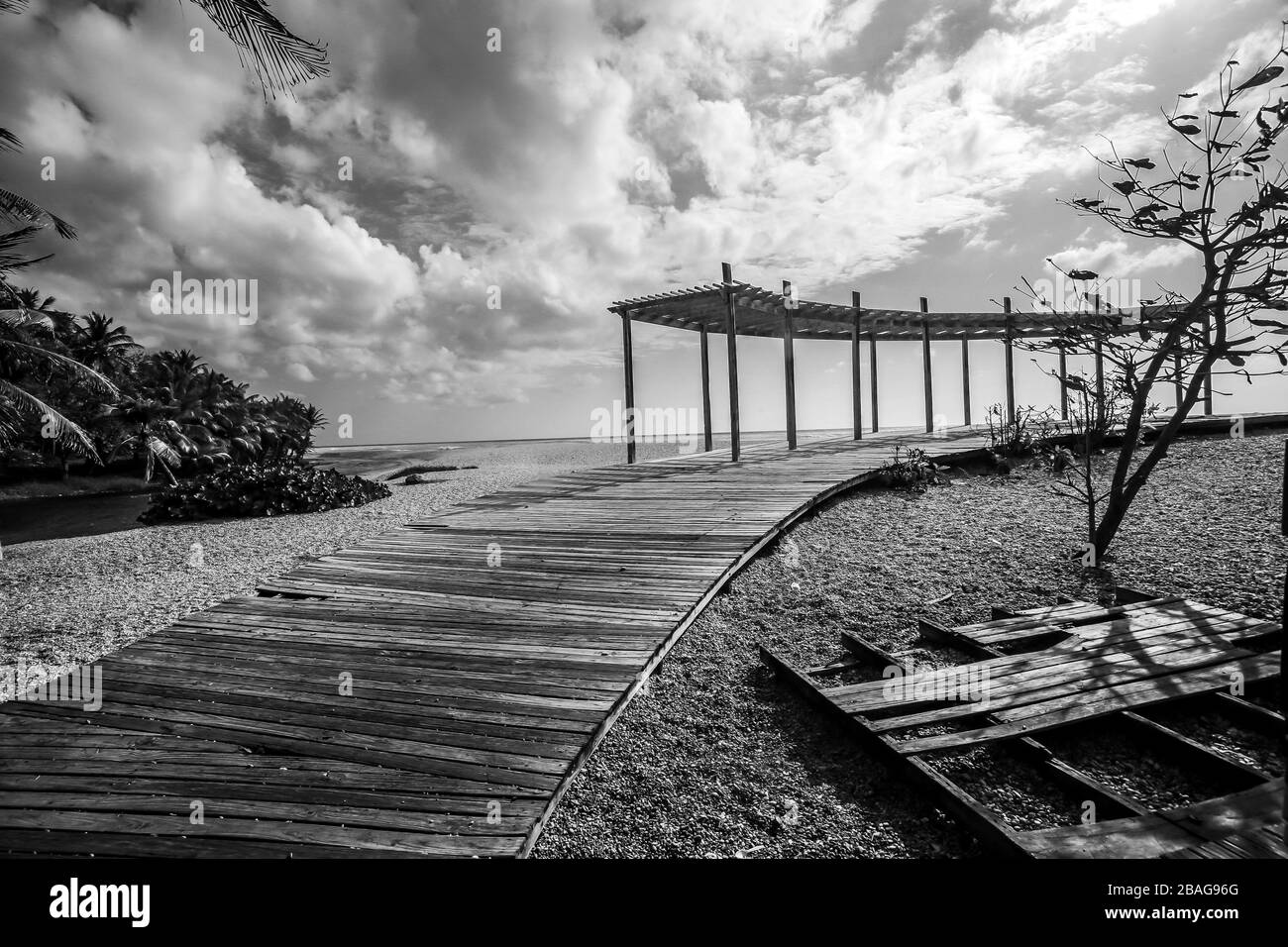 Dramatische Schwarz-Weiß-Seescape der karibischen Küste mit Holzdeck, das sich bis zum Strand erstreckt, und einer Terrasse mit bewölktem Himmel. Stockfoto