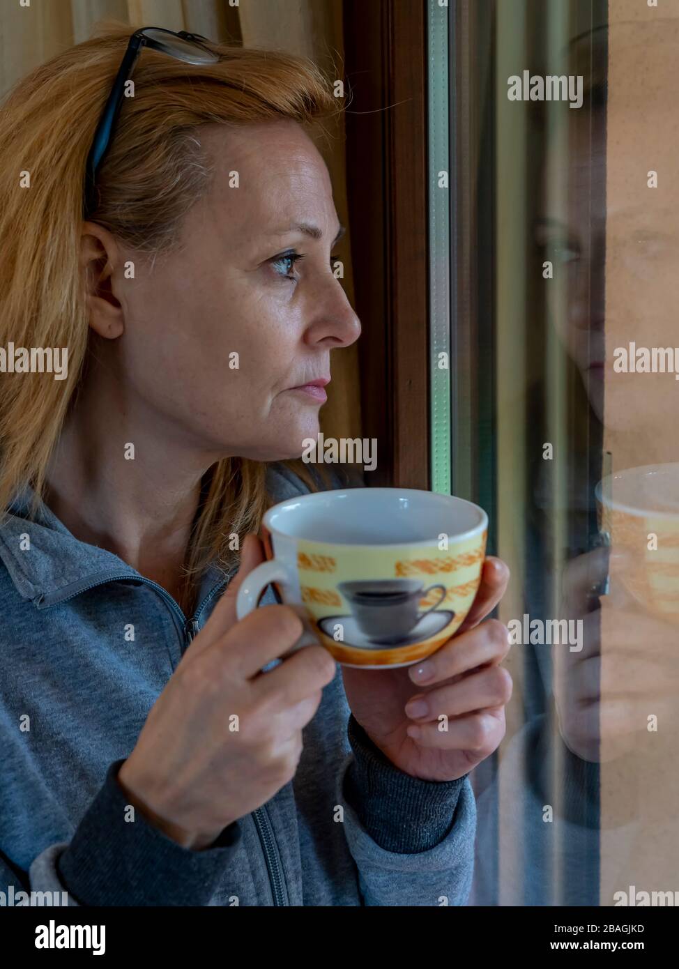 Die unter Quarantäne stehende blonde Frau trinkt von dem Fenster, in dem sie sich widerspiegelt, eine Tasse Tee, der auf die Außenwelt blickt Stockfoto