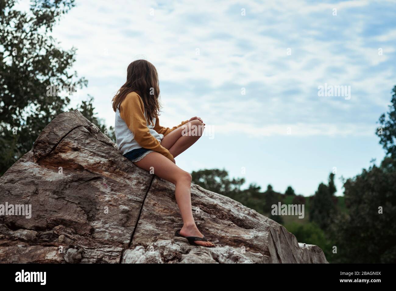 Junges Mädchen 10-12 Jahre alt Blick auf Wolken während auf einem Holzstamm sitzen Stockfoto