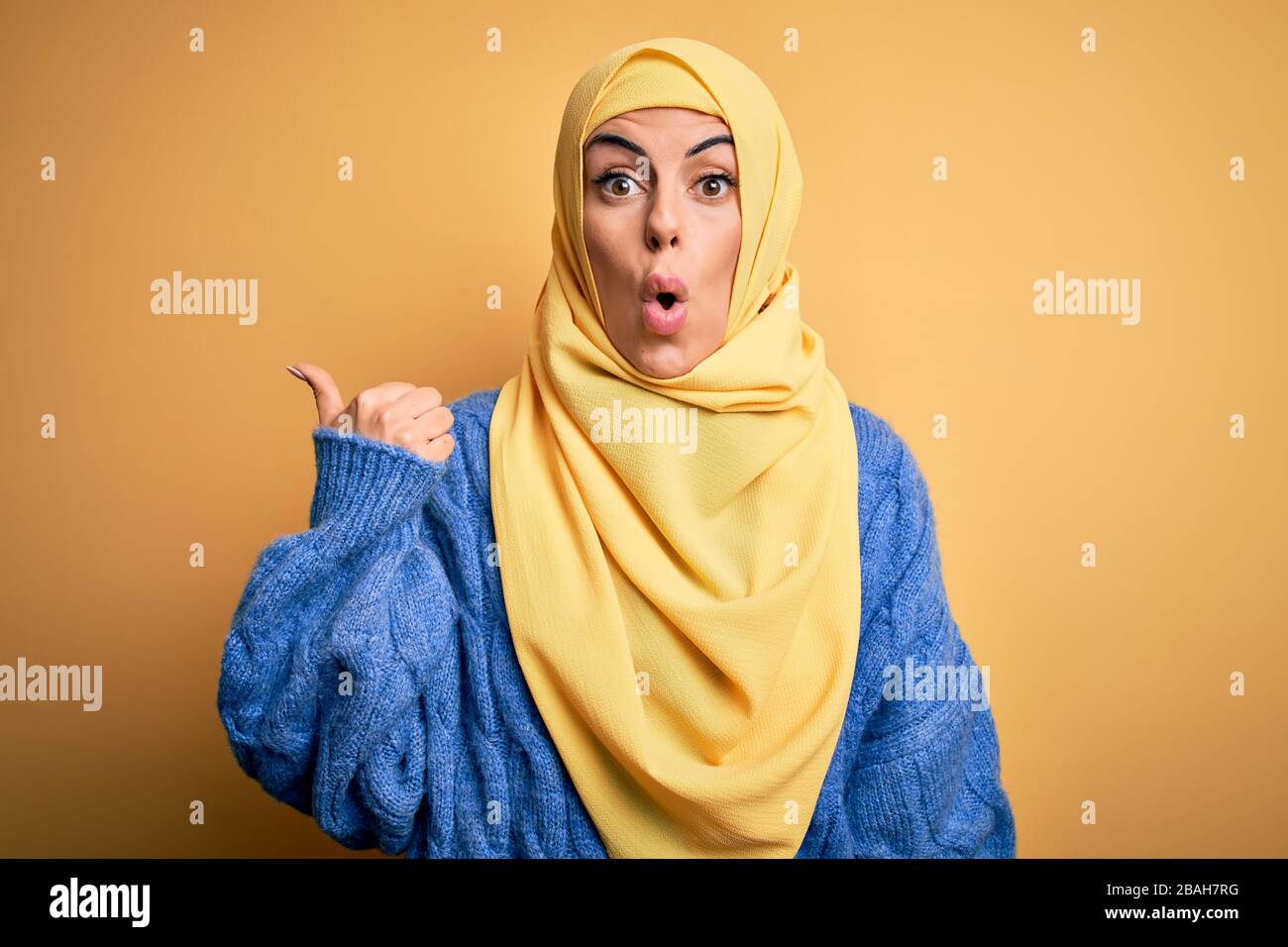 Junge, schöne brünette muslimin, die arabischen Hijab über isoliertem gelbem Hintergrund trägt, überraschte mit dem Handfinger zur Seite, offenen Mund a Stockfoto