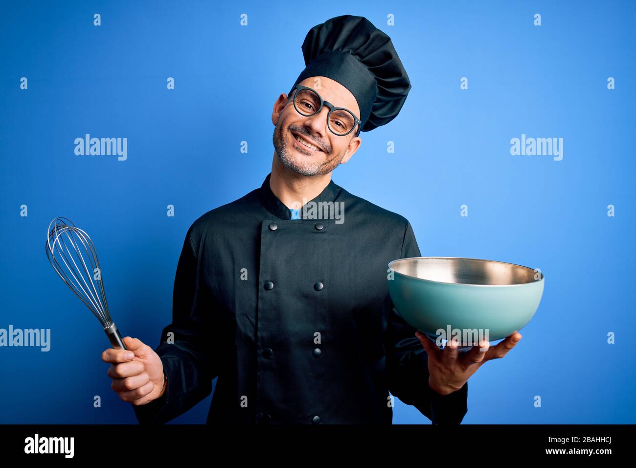 Junger, gutaussehender Küchenchef mit Herduniform und Mischen von Hut mit Schneebesen und Schüssel mit einem fröhlichen Gesicht stehend und lächelnd mit einem selbstbewussten Lächeln Stockfoto