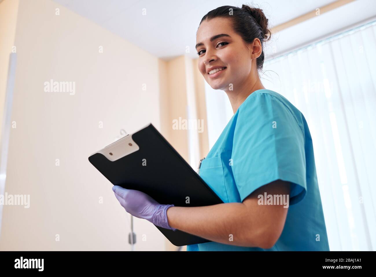 Portrait der lächelnden, attraktiven Krankenschwester in chirurgischen Handschuhen, die sich Notizen auf der medizinischen Karte machen, während sie im Krankenhaus Runden macht Stockfoto