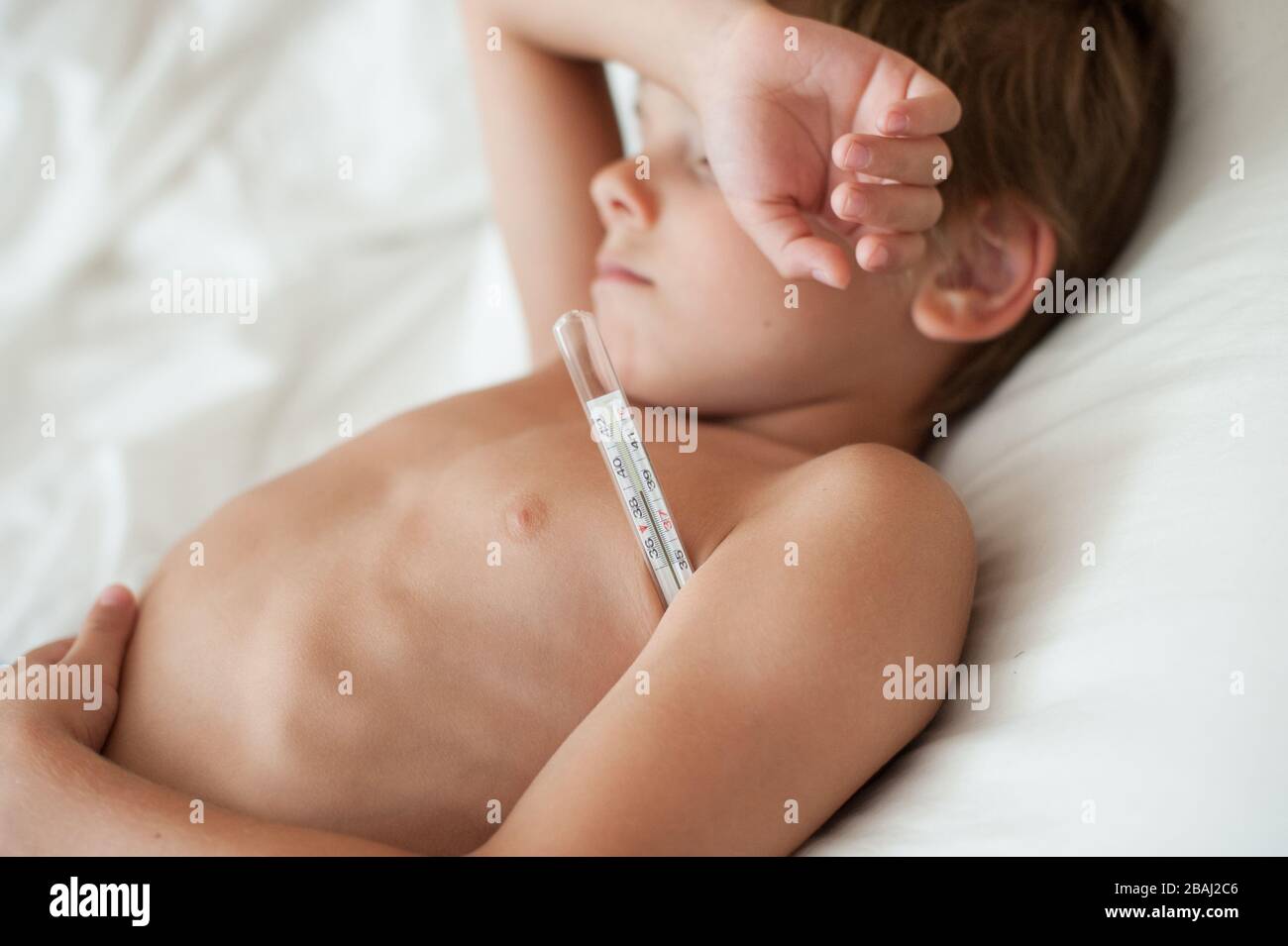 Kranker kleiner Junge mit hoher Temperatur, der im Bett mit Thermometer unter dem Arm während der Kovid-19-Epidemie des Pandemieausbruchs liegt Stockfoto