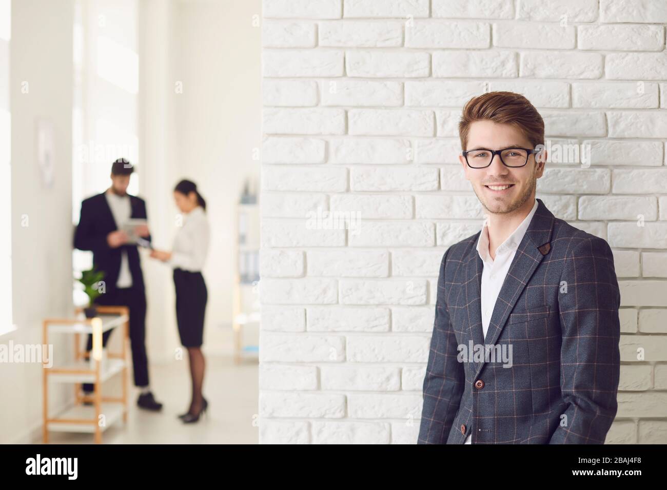 Lächelnder positiver Geschäftsmann in legerer Kleidung, der in einem weißen Büro an einer weißen Wand steht. Stockfoto