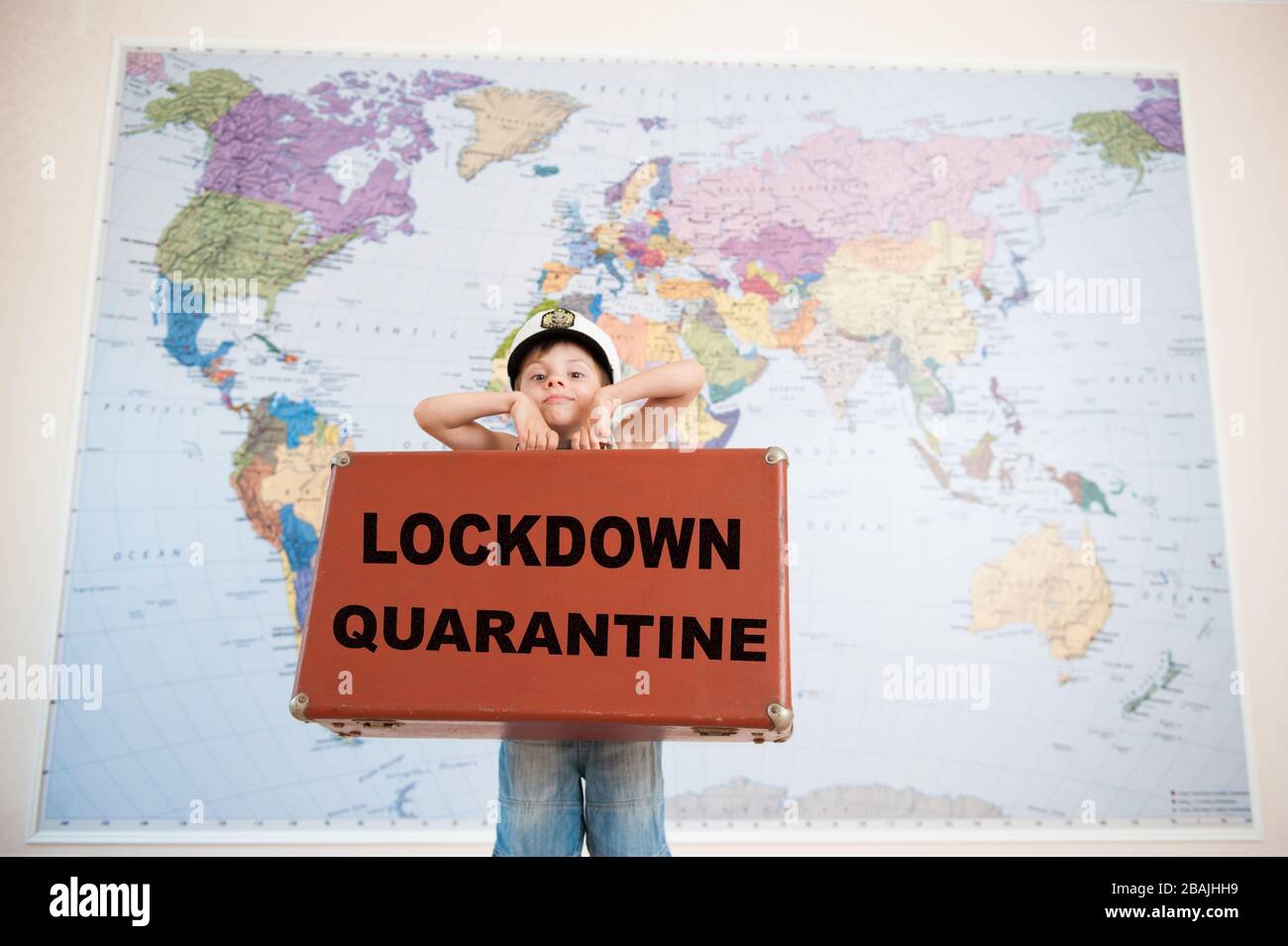 lockdown Quarantäne CANCEL Travel Concept of Little Boy in Kapitän hat Holdingcase mit Slogan auf Weltkarte Hintergrund Stockfoto