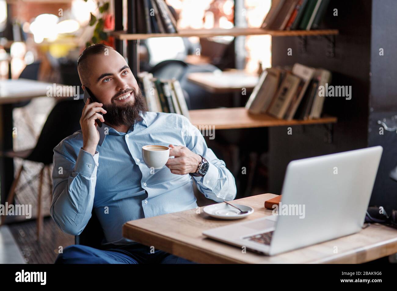 Der junge lächelnde bärtige Geschäftsmann sitzt am Tisch vor dem Computer, spricht auf dem Handy und hält Stift. Freiberufler arbeitet zu Hause. Telefon-Co Stockfoto