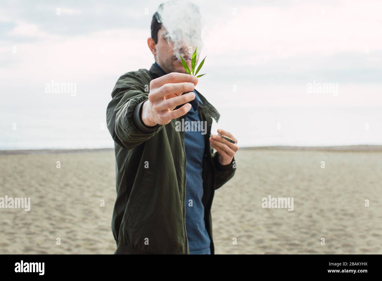 Junger Mann, der ein Marihuanablatt hält und eine Cannabisverbindung am Strand raucht. Hintergrund verwischen. Stockfoto