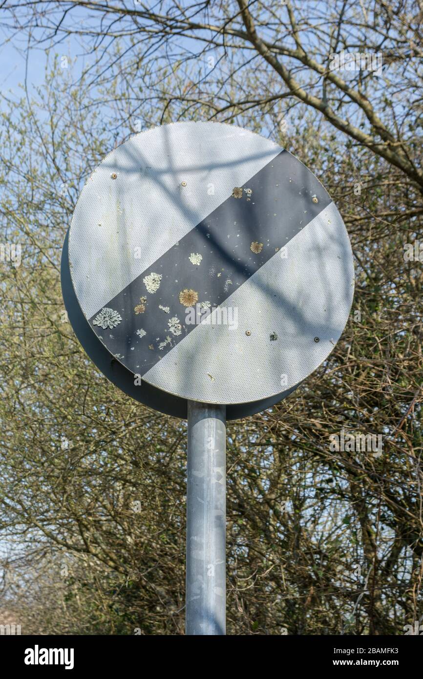 Verblichene und Flechten bedeckte UK National Speed Limit Straßenschild - weißer Kreis mit schwarzer Linie - auf einer ruhigen Landstraße im Frühling Sonnenschein. Stockfoto