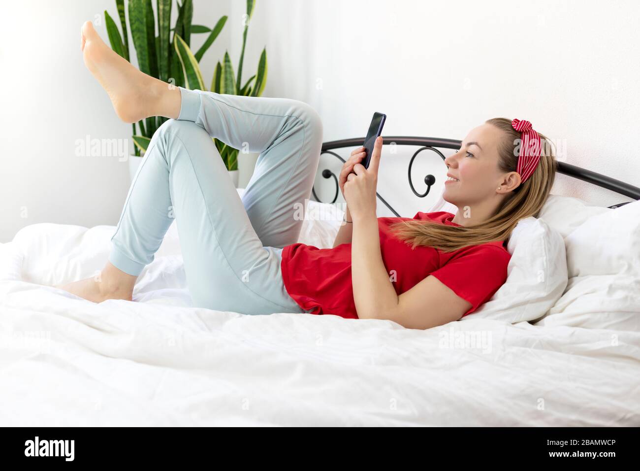 Junge lächelnde Frau mit blonden Haaren in einem roten T-Shirt und bequemen Hosen liegt auf einem weißen Bett mit Smartphone, sie arbeitet, studiert von zu Hause, schreibt A. Stockfoto