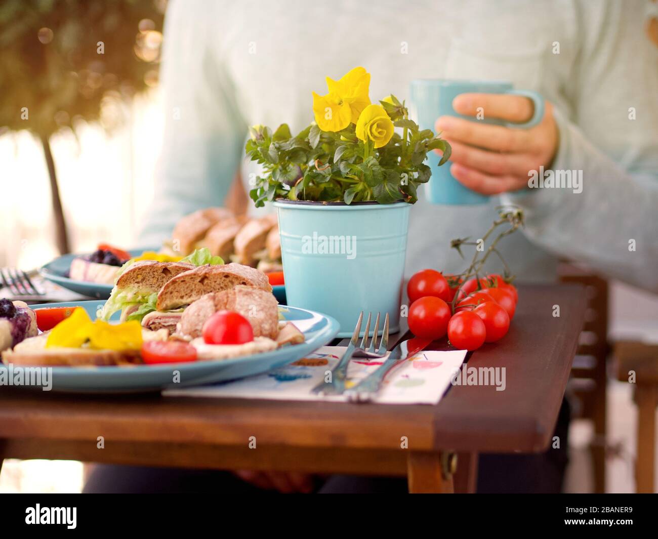 Wunderschönes, farbenfrohes Frühlingsfrühstück mit verschiedenen Speisen auf einem Holztisch mit Blumen an einem sonnigen Tag und einem Mann im Hintergrund. Stockfoto