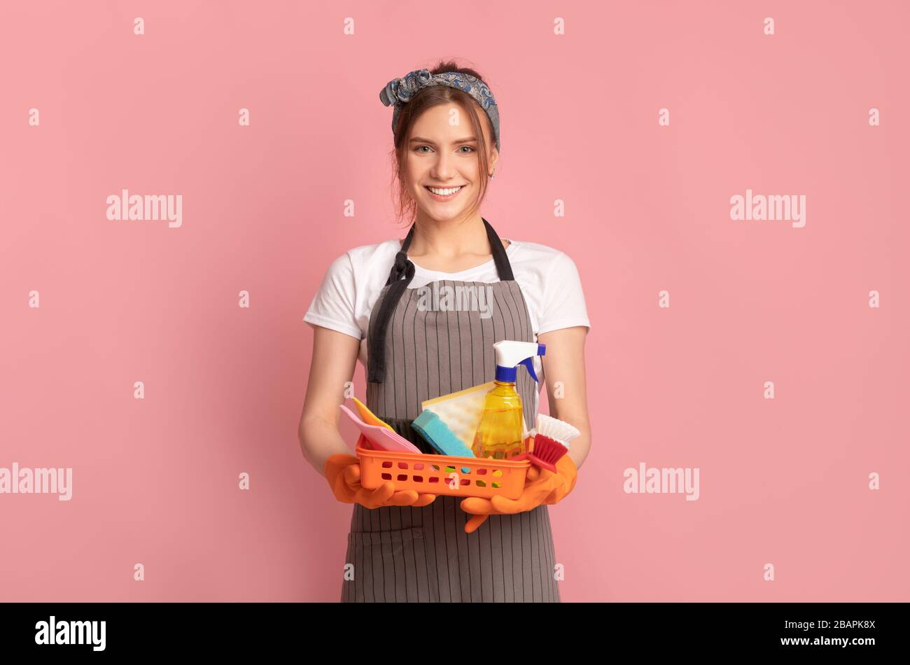 Lächelnde Frau Im Schürze Holding Basket Mit Reinigungszubehör, Pinker Hintergrund Stockfoto