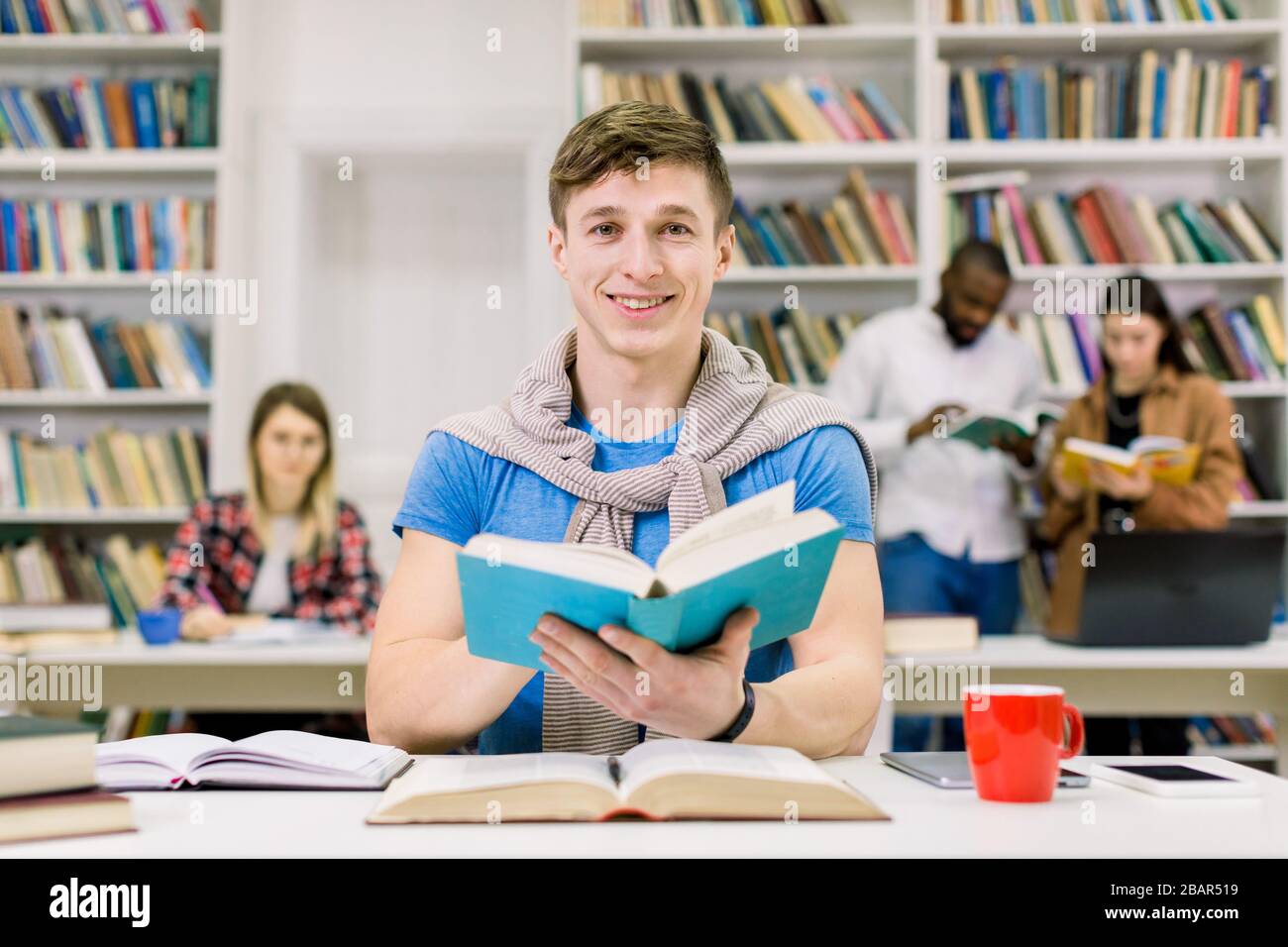 Junge lächelnde Hippie-Studentin, die in der Bibliothek an der Universität studiert. Student schaut mit seiner Kamera und lächelt, genießt es, während er mit seiner in der Bibliothek studiert Stockfoto
