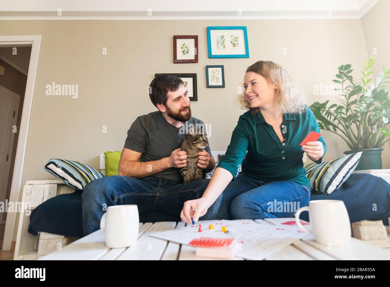 Junges osteuropäisches Paar spielt zu Hause Tischspiel während Corona-Virusquarantäne. Lächeln und sich gegenseitig betrachten. Stockfoto