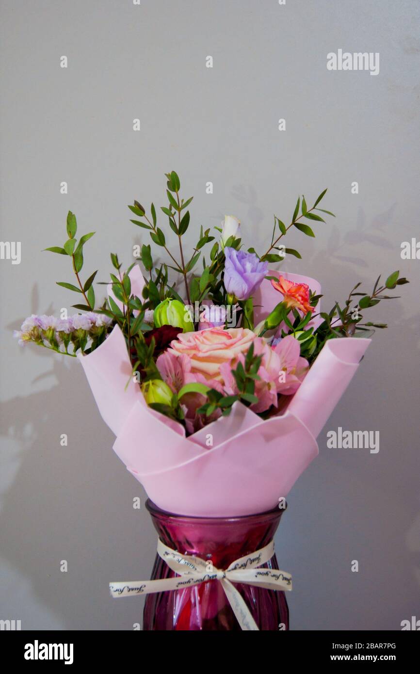 Ein zartes Blumenstrauß verschiedener Blumen in einem hellrosa Wrapper steht in einer weinroten Vase auf grauem Grund. Vertikales Foto. Stockfoto