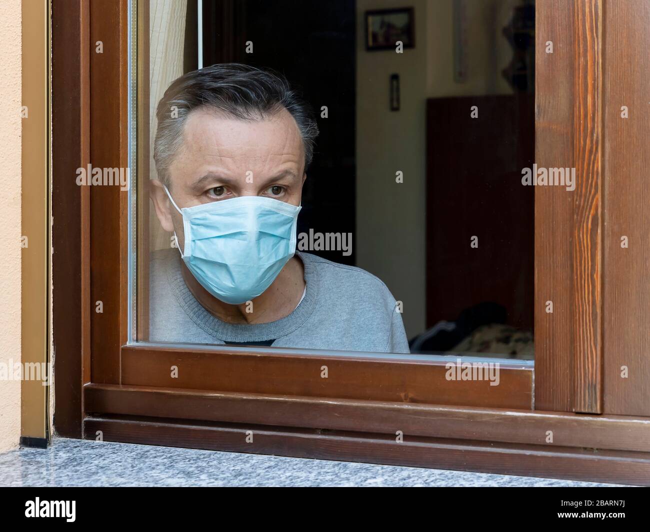 Der italienische Mann mit Schutzmaske, der aufgrund der Coronavirus Covid-19 zu Hause bleiben musste, blickt trostlose auf das Fenster des Hauses Stockfoto
