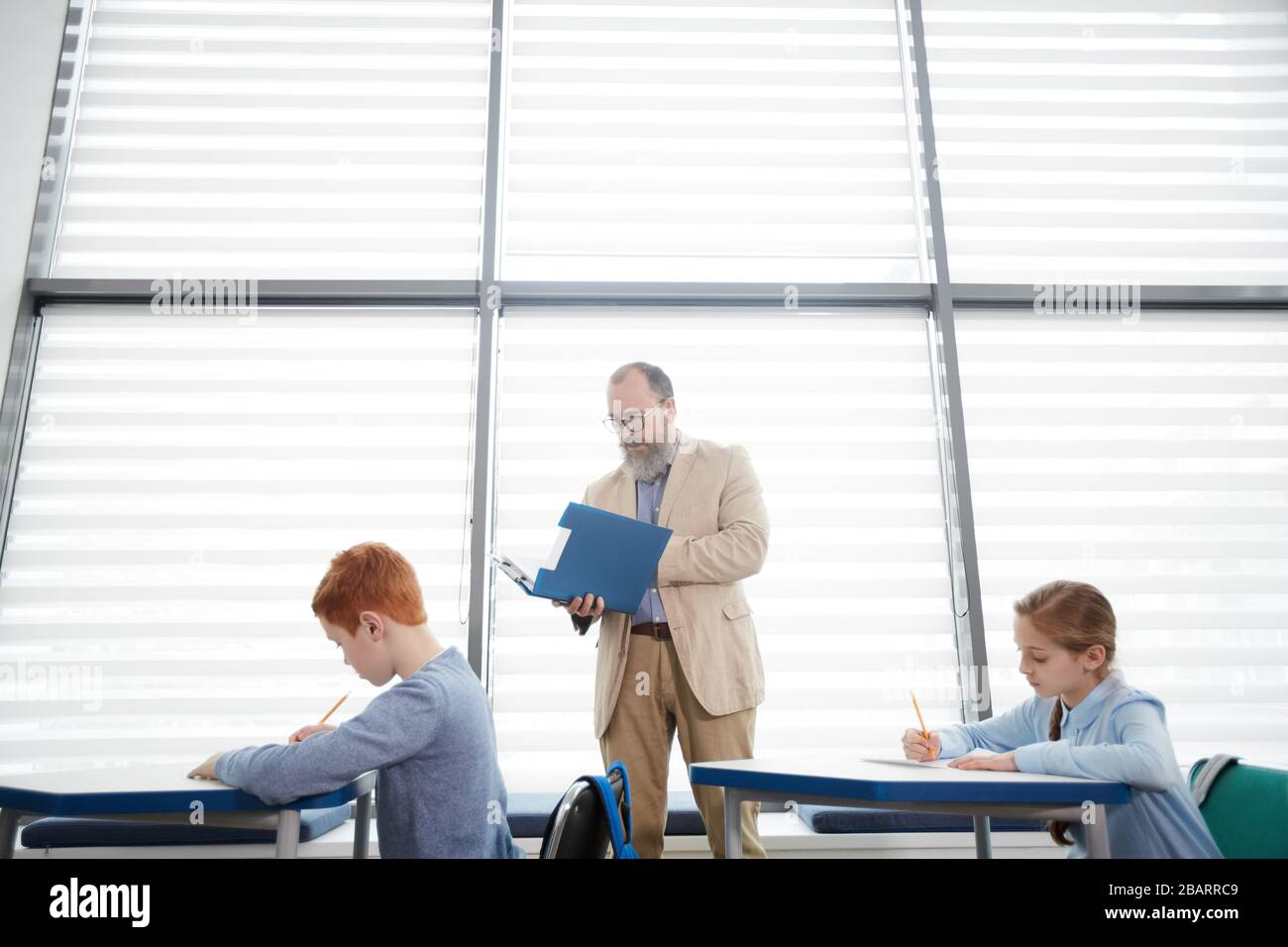 Seitenansicht bei reifer bärtiger Lehrerin, die Kinder beobachtet, die im Schulunterricht Tests machen, Kopierraum Stockfoto