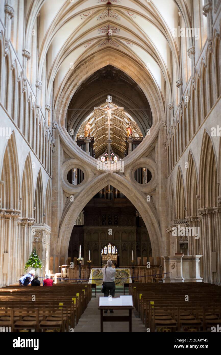 Doppelter Wishbone Arch und Vorderseite der Wells Cathedral: Der zusätzliche kultige Truss bewahrt die Integrität des alten Gebäudes. Oben ein Kruzifix, darunter ein Flügelaltar. Ein Fotograf schießt auf den vorderen Domaltar, während andere Touristen hinschauen. Stockfoto