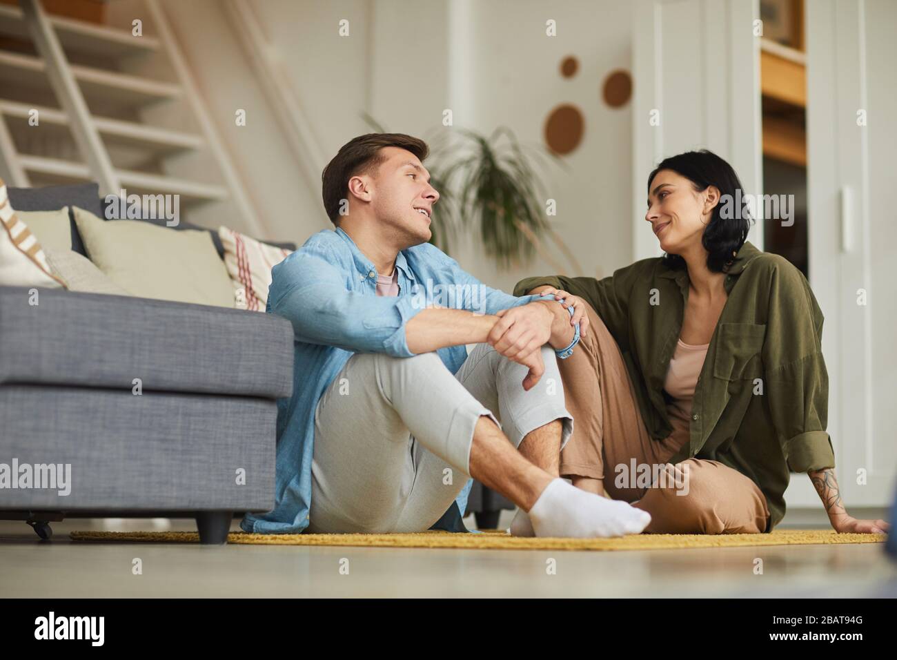 Warmes, langes Porträt eines modernen jungen Paares, das sich aufrichtig miteinander spricht, während es im gemütlichen Innenbereich auf dem Boden sitzt und Platz für Kopien bietet Stockfoto