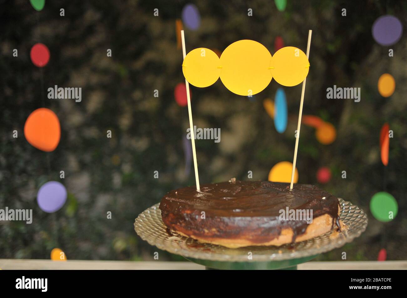 Einfacher, handgefertigter Geburtstagstorte aus Schokolade in einer dekorativen Platte mit gelben Kreisen, die ihn schmücken. Stockfoto