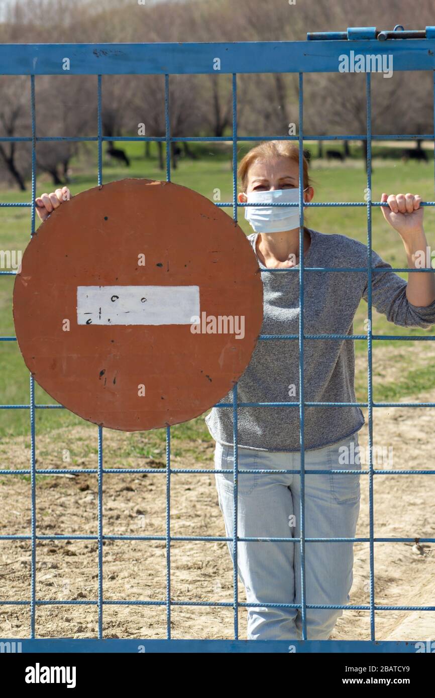 Kaukasische Frauen stehen in Schutzmaske hinter verschlossenem Tor mit rotem Stoppschild. In Quarantäne für Coronavirus Kovid-19 . Das Konzept "zu Hause bleiben" Stockfoto
