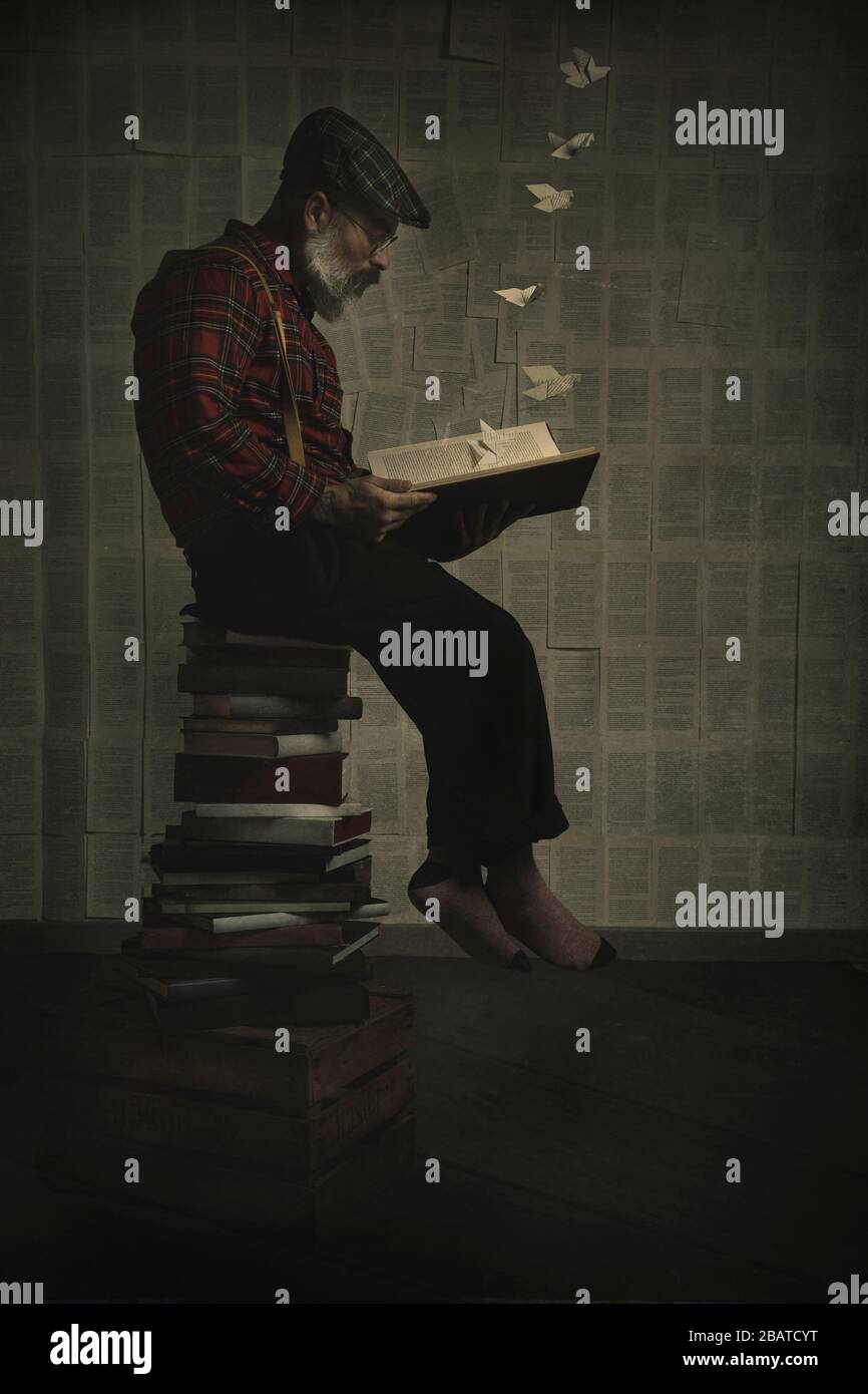 Der Mann sitzt auf einem Stapel von Büchern, die ein Buch lesen, das der Phantasie ein surreales Bild lässt. Vertikales Bild Stockfoto