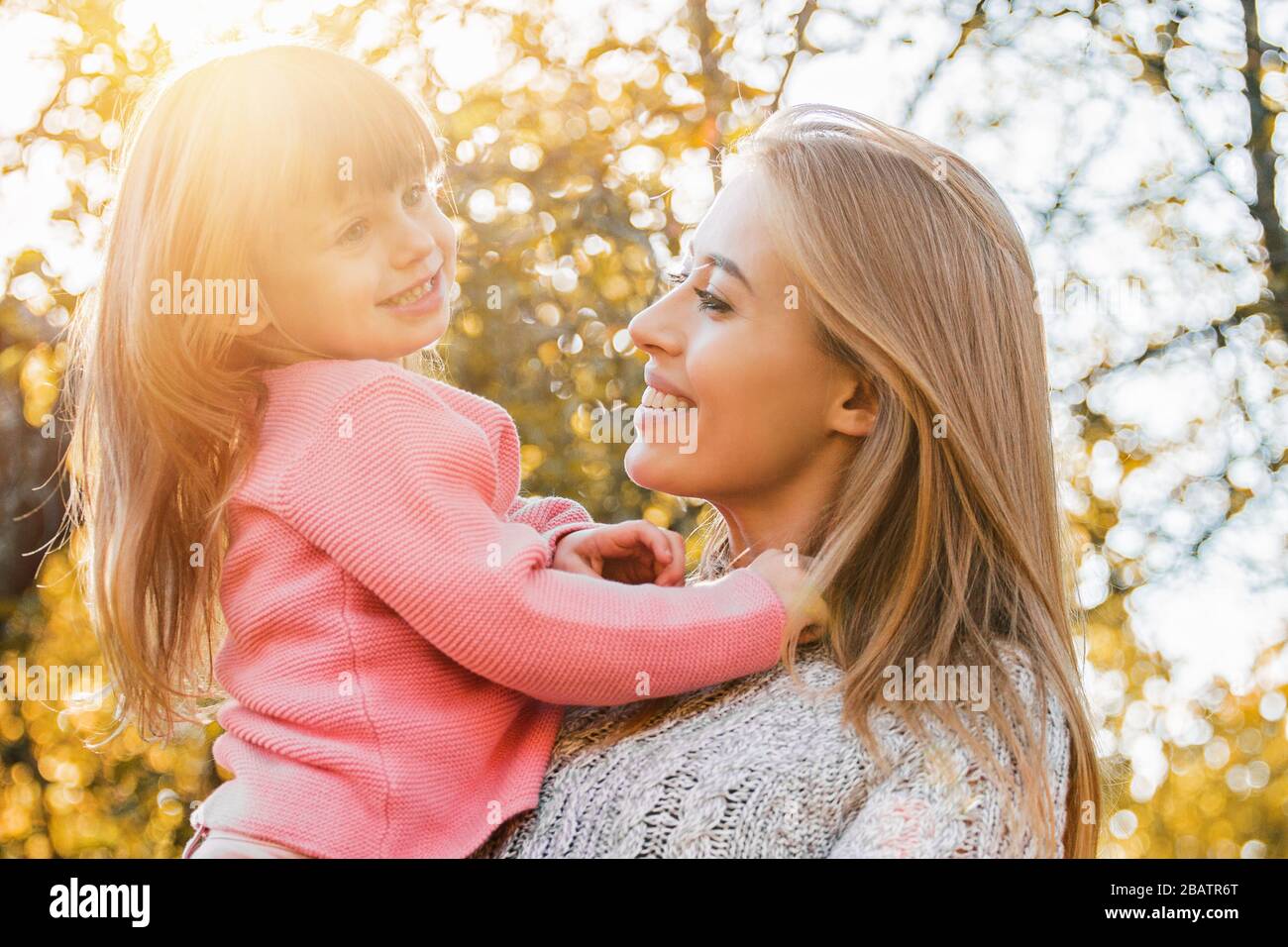 Fröhliche junge Familie, die sich im Herbstpark umarmt. Fröhliche junge Mutter umarmt ihre schöne kleine Tochter, während sie im Freien steht. Fröhliche Frau hält Stockfoto