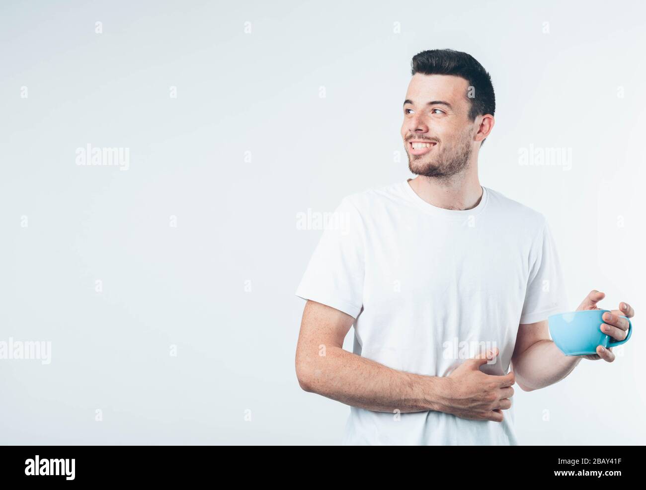 Junger brünette Mann mit Bart in weißem T-Shirt. Guy lächelt eine Hand auf seinem Bauch, die zweite hält den blauen Becher. Isoliert über hellem Hintergrund. Stockfoto