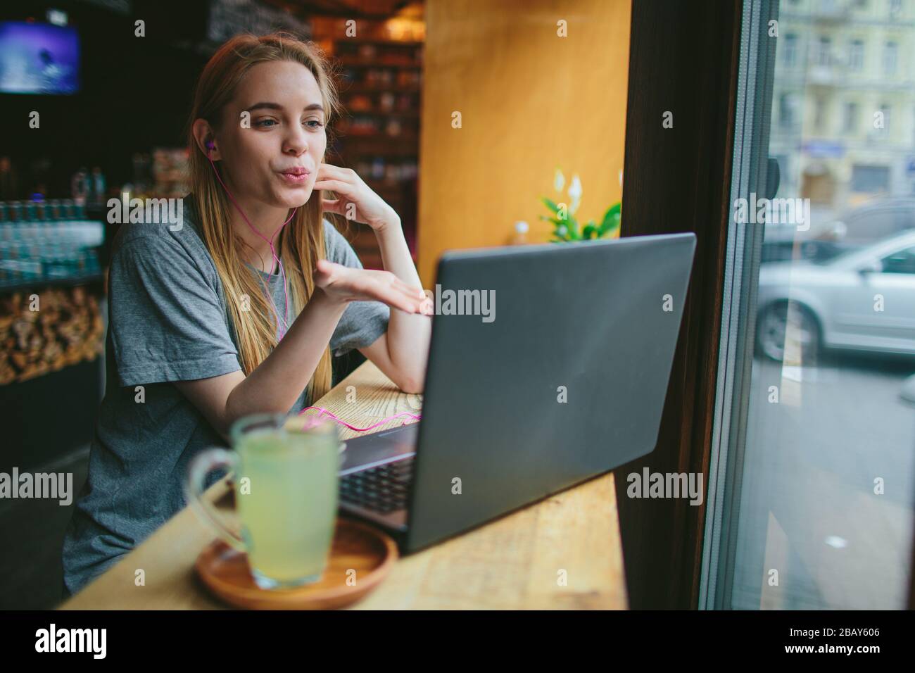 Die junge Blonde in einem Café arbeitet mit einem Laptop. Sie spricht über Skype und bläst einen Kuss. Auf dem Tisch steht eine Tasse grünen Tee. Stockfoto