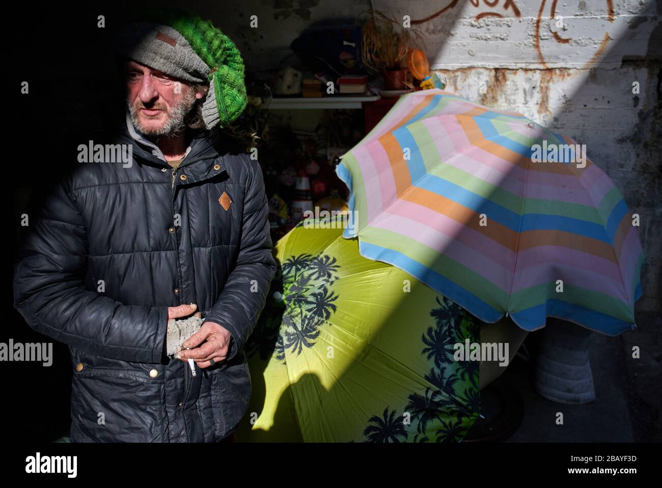 München, Deutschland. März 2020. "Gogo" steht an seinem Schlafplatz unter einer Eisenbahnbrücke, wo er sich mit Sonnenschirmen vor Wind schützt. Obdachlose sind besonders von der Corona-Pandemie betroffen. Das Hilfesystem zerfällt zunehmend, gleichzeitig müssen die oft immungeschwächten Menschen vor allem eine Infektion fürchten. Credit: Bernd Thissen / dpa / Alamy Live News Stockfoto