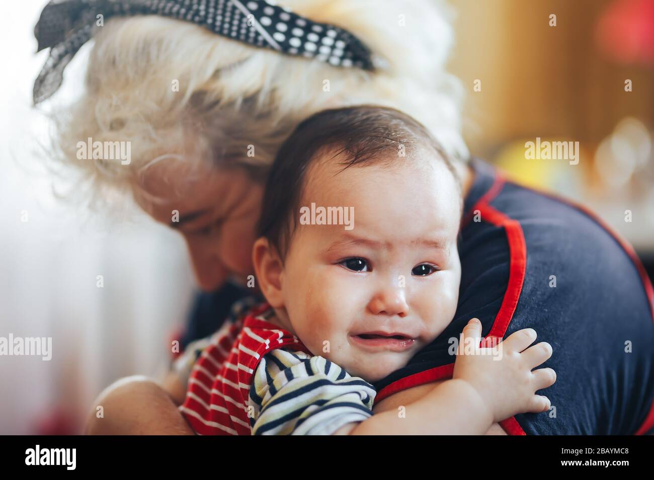 Großmutter, die weinendes Mädchen im Säuglingsalter mit Blick auf die Kamera umschlindet Stockfoto