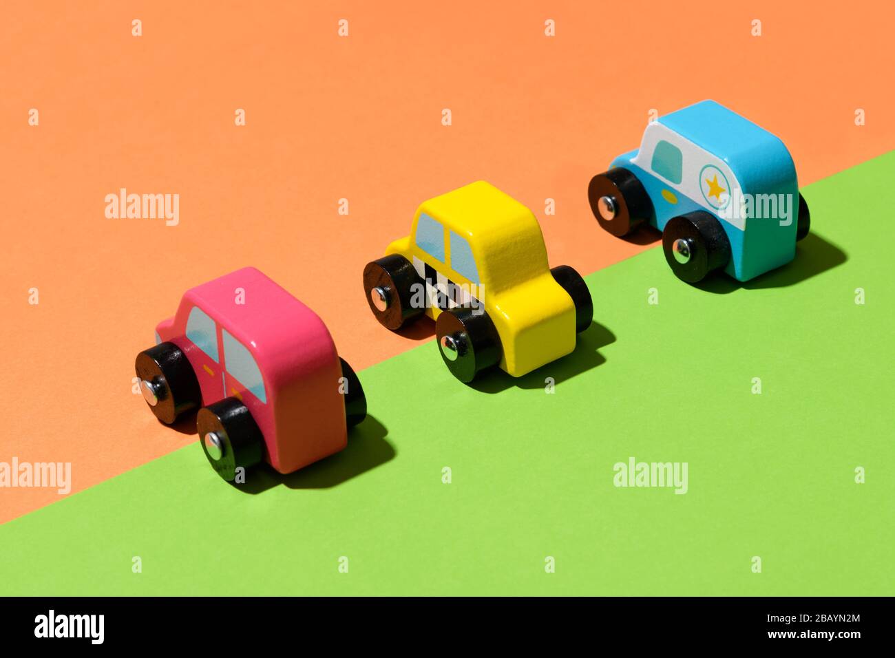 Drei bunte, rustikale, handgefertigte Spielzeugautos aus Holz auf einem geteilten grünen und orangefarbenen Hintergrund, die entlang der diagonalen Farbverteilung aufgereiht sind Stockfoto