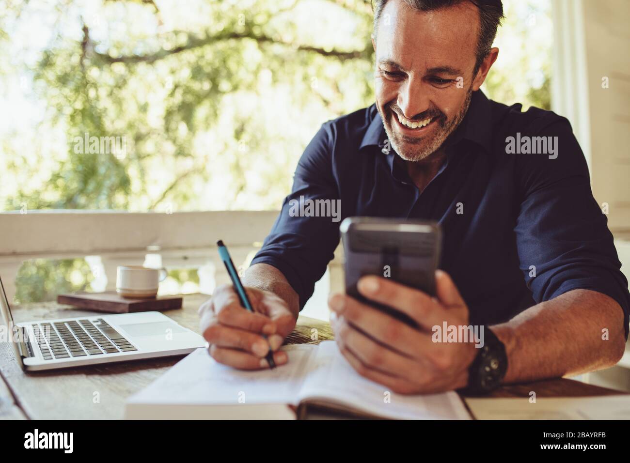 Lächelnder Geschäftsleute sitzt am Tisch im Café mit Laptop, während er Smartphone benutzt und in sein Tagebuch schreibt. Stockfoto