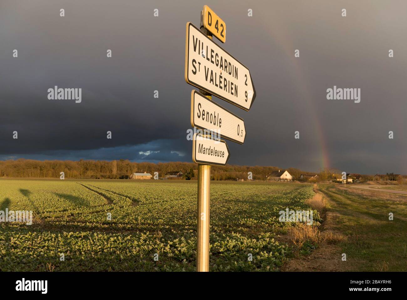 Französischer Wegweiser auf der D42 bei Villegardin mit Regenbogen in Feldern gegen dunklen Himmel Stockfoto