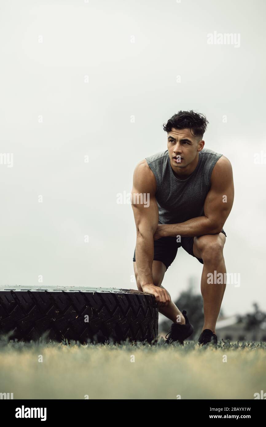 Passen Sie jungen Mann, der im Freien ein Flip-Workout für Reifen macht. Muskelsportler, der eine Pause vom Cross-Training auf einem Feld nimmt. Stockfoto
