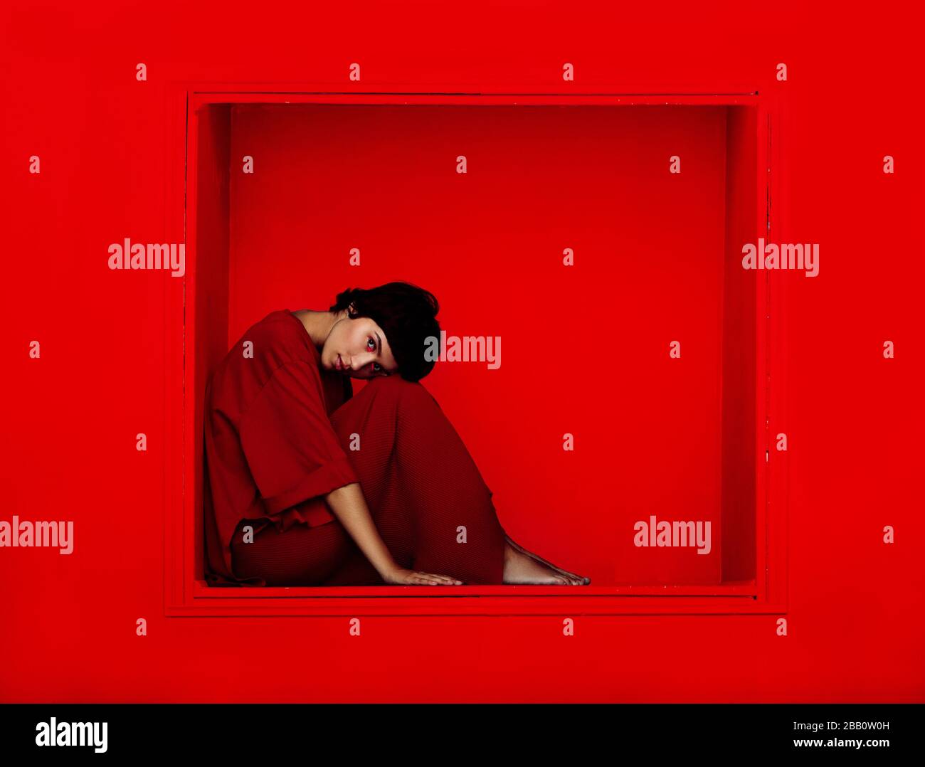 Modefoto der jungen schönen Frau, die in einer Nische auf rotem Hintergrund sitzt. Selbstisolierungskonzept. Stockfoto