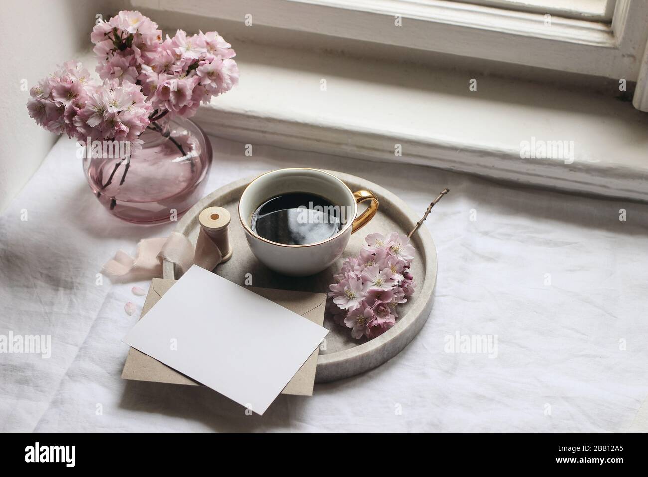 Frühlingsszene im Stillleben. Grußkarten Mockups, Umschlag, Marmortablett, Tasse Kaffee, Vintage feminin gestyltes Foto. Blumenapposition mit rosafarbenem sak Stockfoto