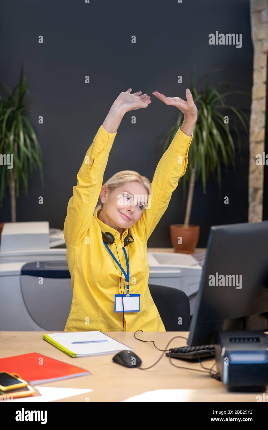 Müde blonde Frau sitzt in Kopfhörern am Schreibtisch und streckt sich aus Stockfoto