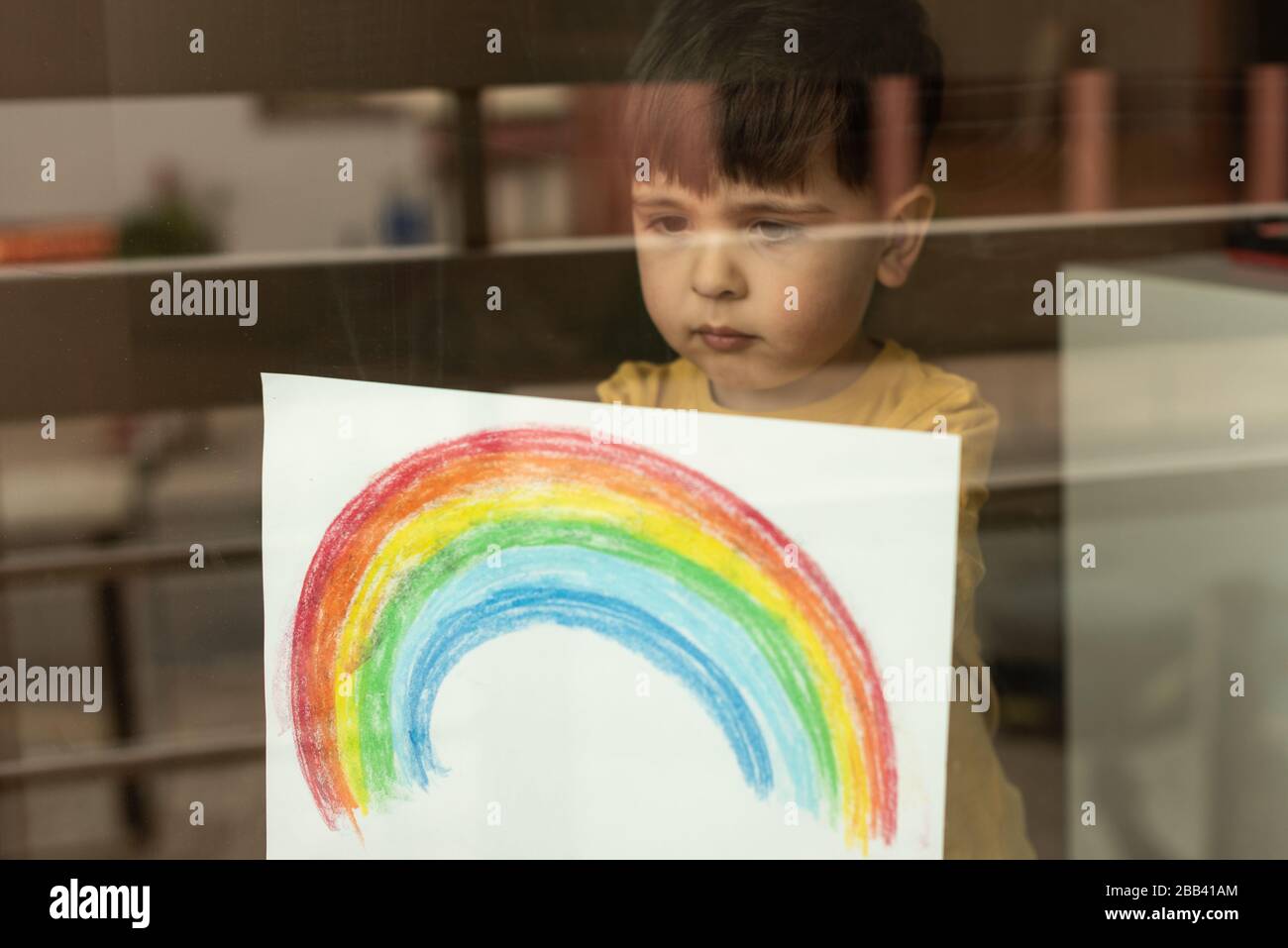 Inspirierendes kleines Kind, das eine Zeichnung eines Regenbogens durch das Fenster hält Stockfoto