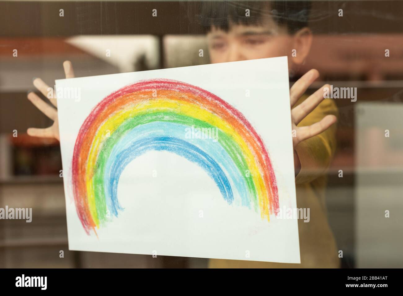 Inspirierendes kleines Kind, das eine Zeichnung eines Regenbogens durch das Fenster hält Stockfoto