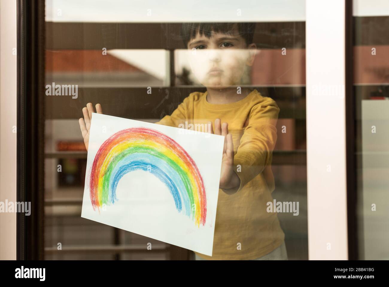 Inspirierendes kleines Kind, das eine Zeichnung eines Regenbogens durch das Fenster hält Stockfoto