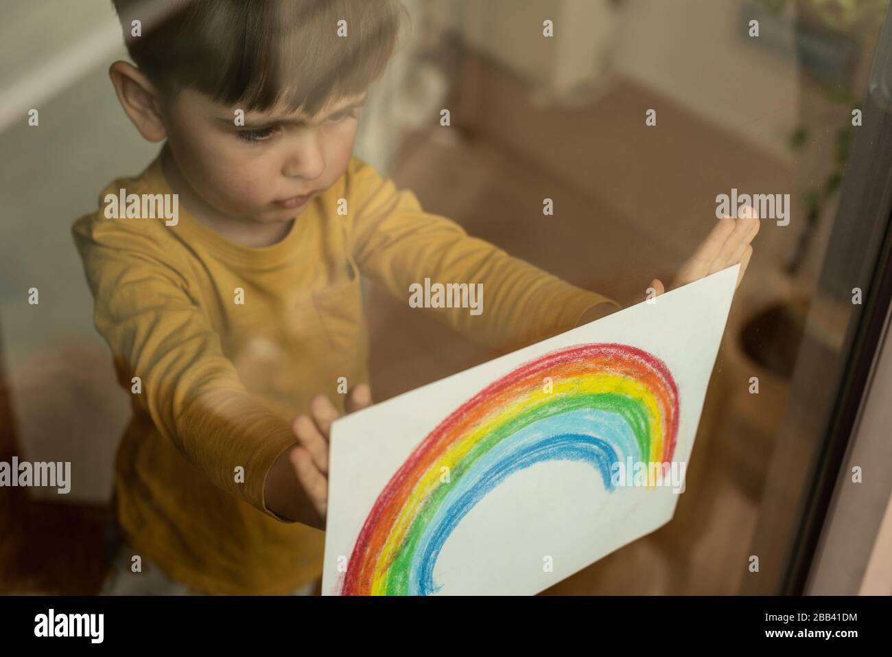 Inspirierendes kleines Kind, das eine Zeichnung eines Regenbogens durch das Fenster hält Stockfoto
