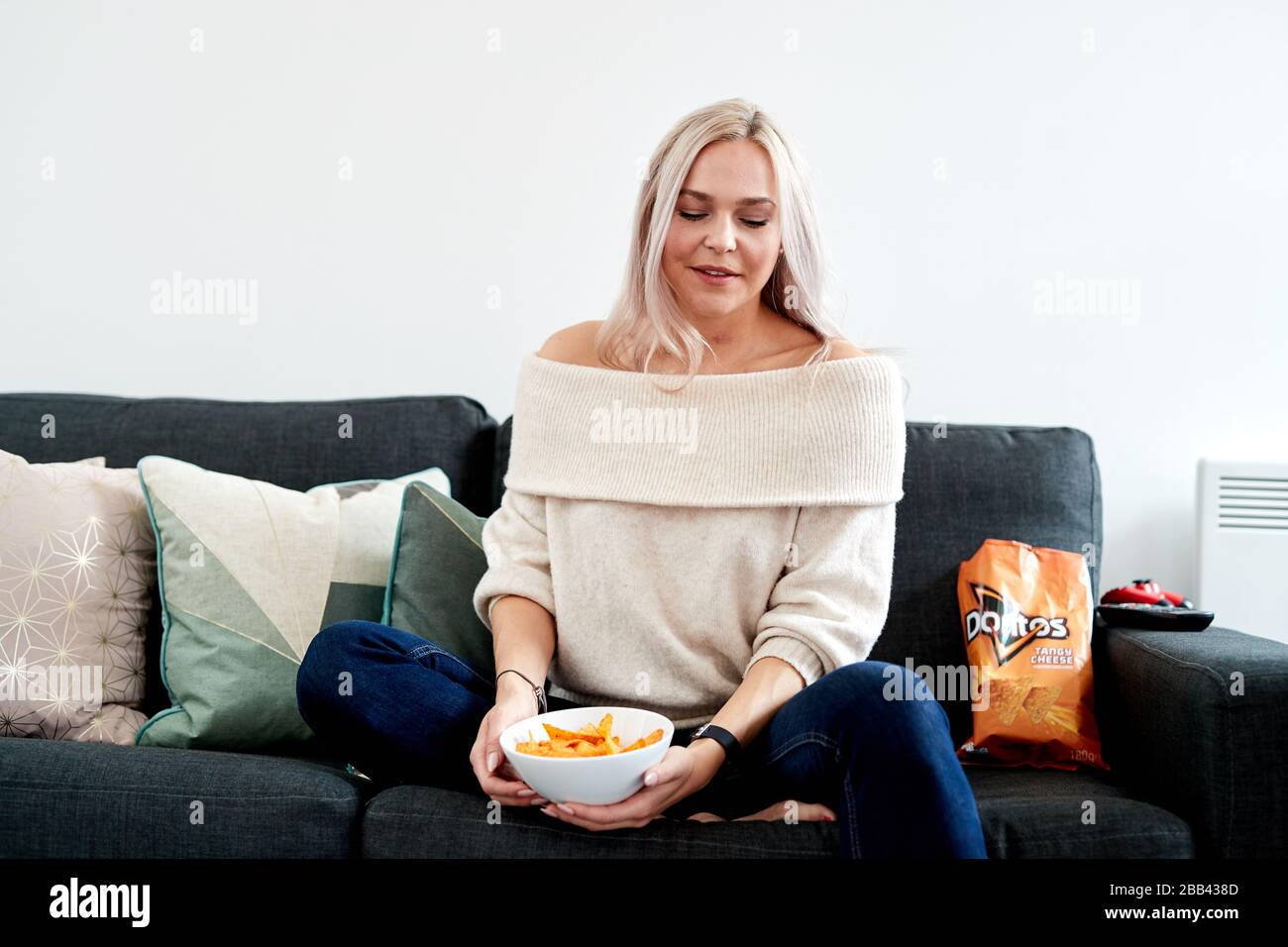 Eine einzelne blonde kaukasische Frau, die auf einem Sofa sitzt und eine weiße Schüssel mit tangiertem Käse Doritos hat Stockfoto