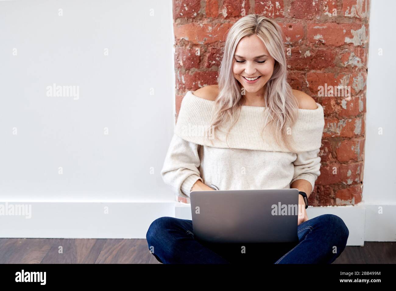 Ein einzelnes, blondes, kaukasisches Weibchen sitzt auf dem dunklen Laminatboden und blickt auf einen Laptop Stockfoto