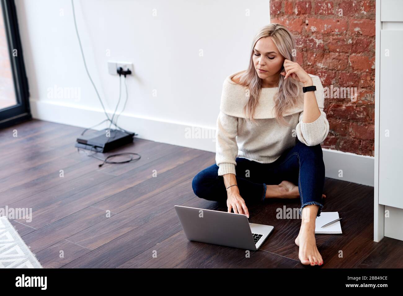 Ein einzelnes, blondes, kaukasisches Weibchen sitzt auf dem dunklen Laminatboden und blickt auf einen Laptop Stockfoto