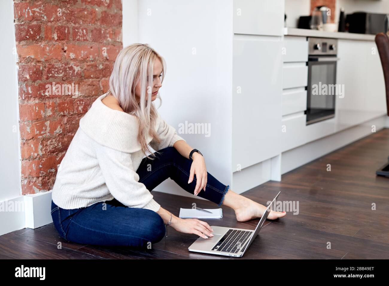 Ein einzelnes, blondes, kaukasisches Weibchen sitzt auf dem dunklen Laminatboden und blickt auf einen Laptop Stockfoto