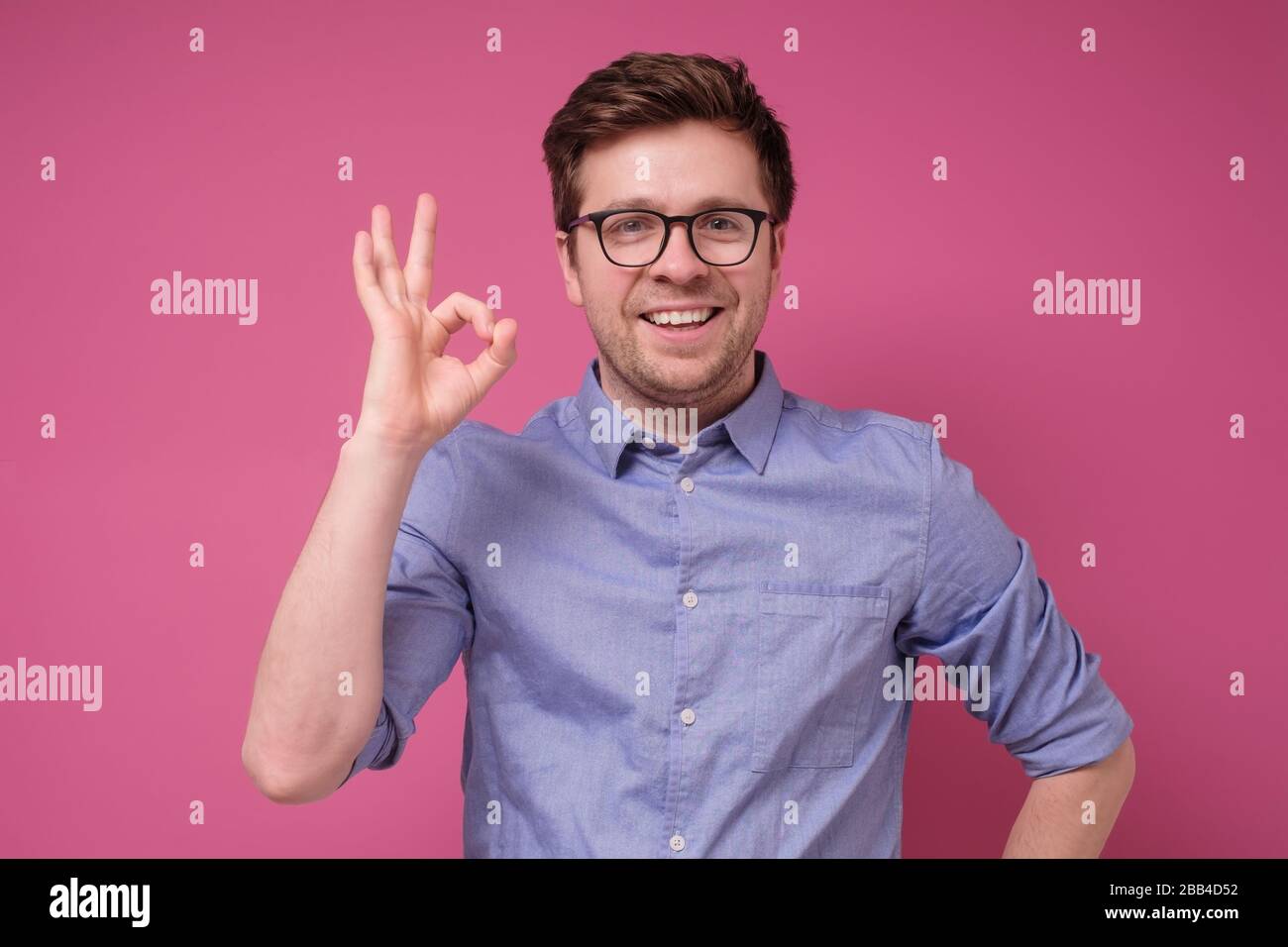 Alles ist in Ordnung. Fröhlicher junger Mann im Hemd, der ein gutes Zeichen gestikuliert und lächelt, während er vor rosafarbenem Hintergrund steht Stockfoto