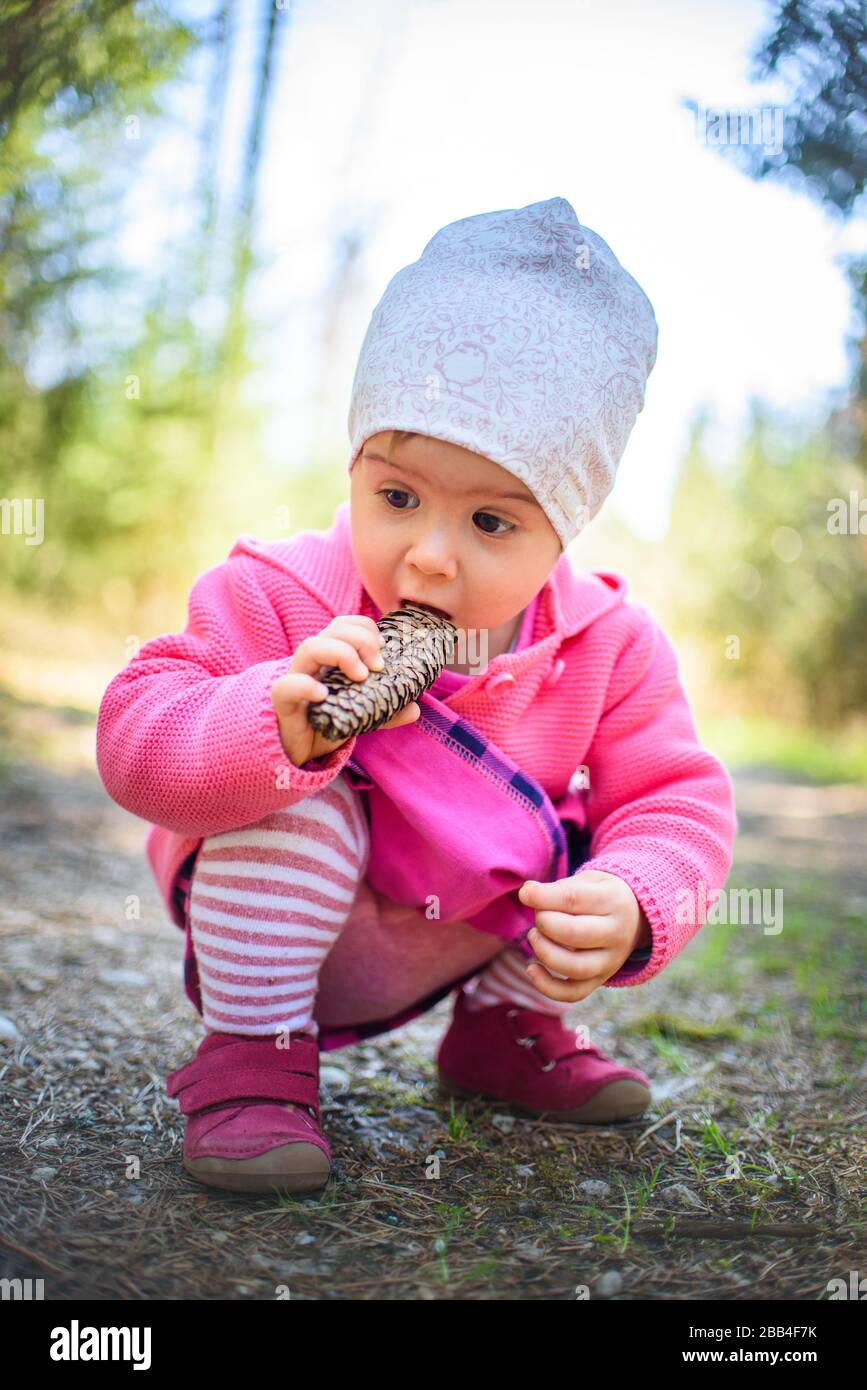 Adliges Baby, das sich auf dem Waldweg in der Natur duckt. Porträt eines 1-jährigen Mädchens im Wald mit Kiefernkegel im Monat. Stockfoto